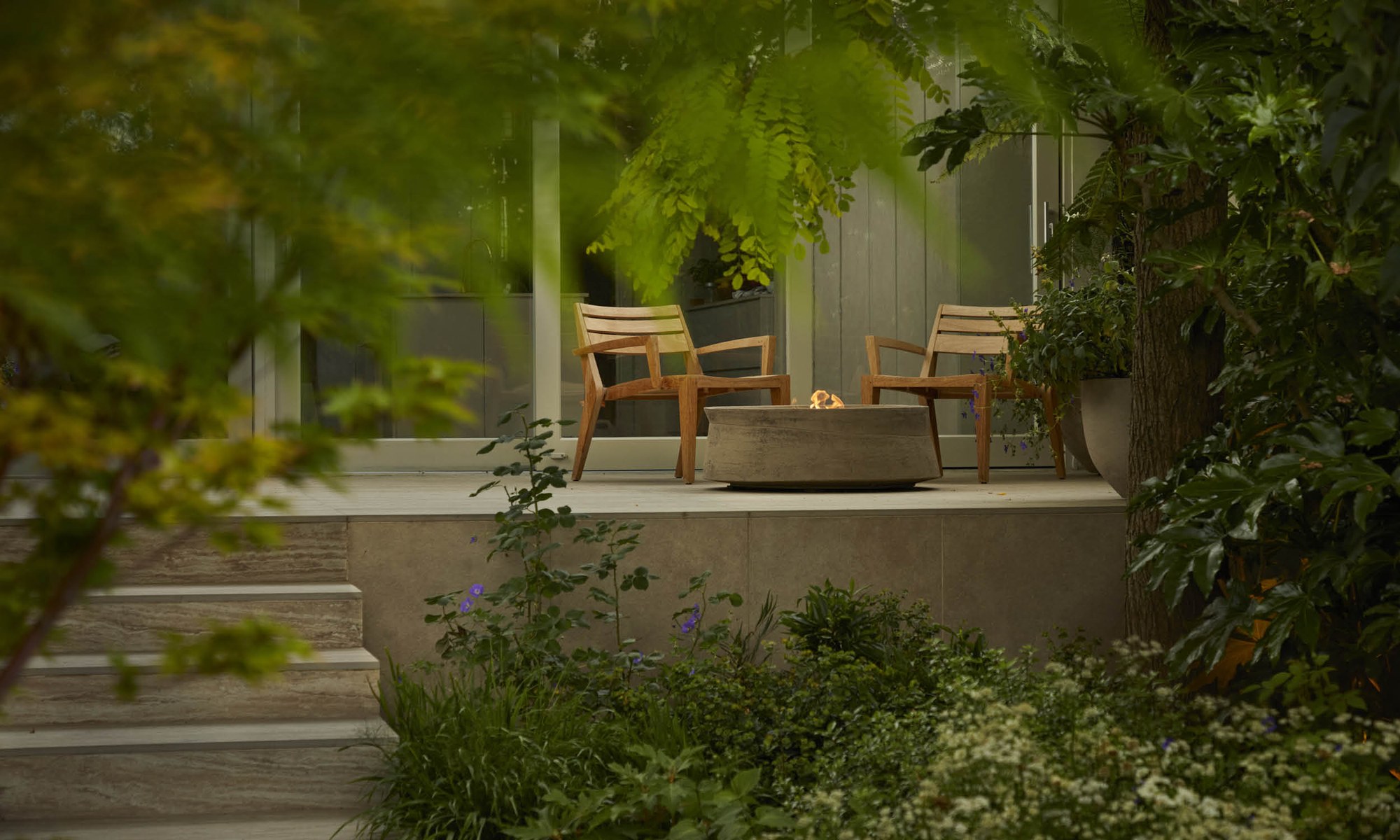 Shaded seating area with a firepit in a Shoots & Leaves garden, surrounded by ferns and trees